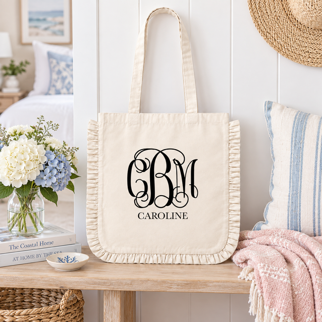 A cream canvas tote bag with ruffled trim and long handles, featuring a black monogram 'CBM' and the name 'CAROLINE' printed on the front. The bag is styled on a wooden table in a bright, coastal-themed bedroom setting with a pillow, blanket, flowers, and a straw hat.
