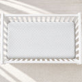 A white crib with a fitted sheet featuring a gray and white chevron pattern, shown from a top-down view on a light textured rug.