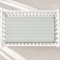 A top-down view of a white wooden crib with a fitted sheet featuring horizontal sage green and white stripes, set against a light beige textured rug with soft window shadow patterns.