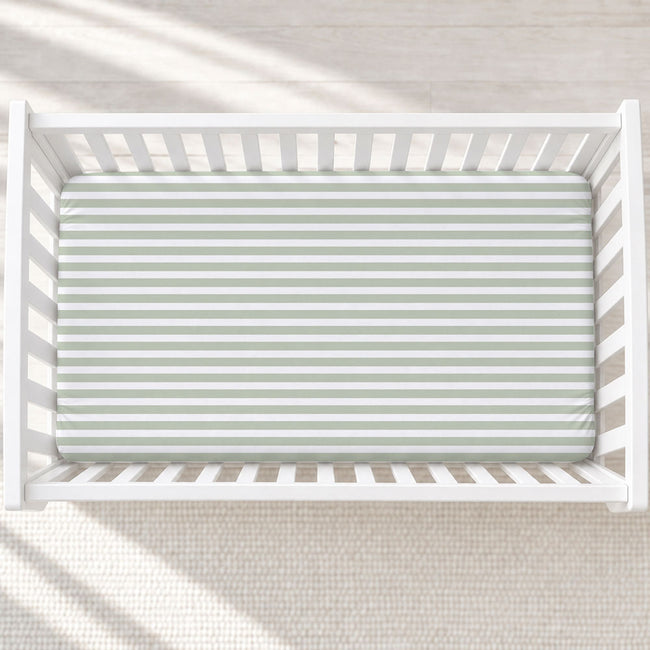 A top-down view of a white wooden crib with a fitted sheet featuring horizontal sage green and white stripes, set against a light beige textured rug with soft window shadow patterns.