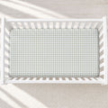 A top-down view of a white wooden crib with a fitted sheet featuring a light sage and white houndstooth pattern, set against a neutral textured rug with soft diagonal shadows.