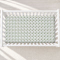 A top-down view of a white wooden crib with a fitted sheet featuring a repeating green and white triangle pattern, set on a light textured rug.