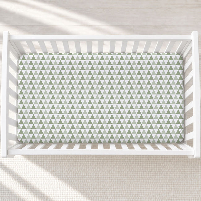 A top-down view of a white wooden crib with a fitted sheet featuring a repeating green and white triangle pattern, set on a light textured rug.