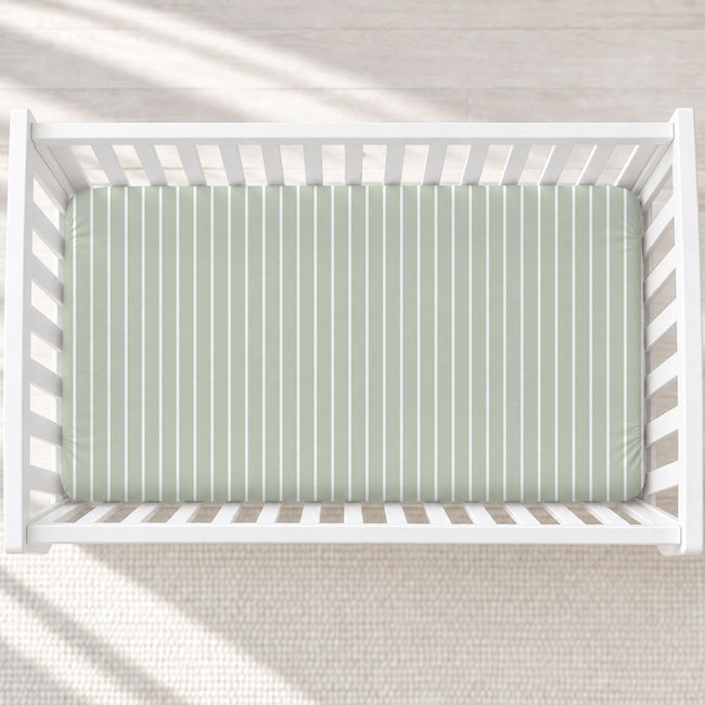 A top-down view of a white wooden crib with a sage green and white vertically striped mattress cover, set against a light beige textured rug with soft diagonal shadows.