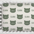A crib bedding set featuring a repeating pattern of dark green cat faces and the name 'ELARA' printed on a warm off-white background, shown inside a white wooden crib.