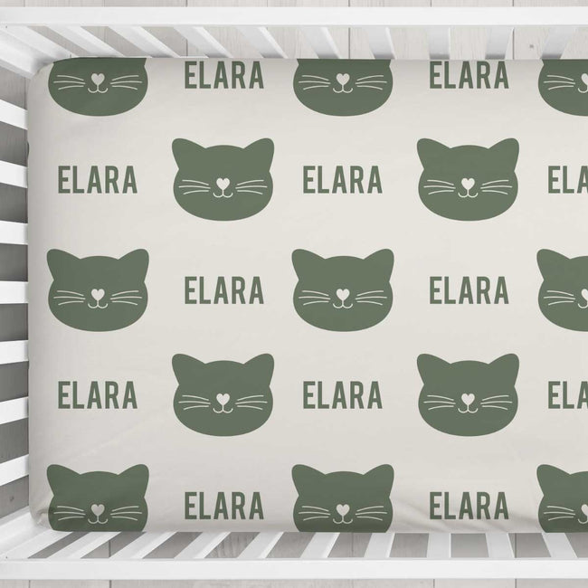 A crib bedding set featuring a repeating pattern of dark green cat faces and the name 'ELARA' printed on a warm off-white background, shown inside a white wooden crib.