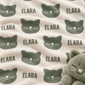 A close-up of a soft fleece blanket with a repeating pattern of dark green cat faces and the word 'ELARA' on a warm cream background. A plush gray cat toy is partially visible in the lower right corner.