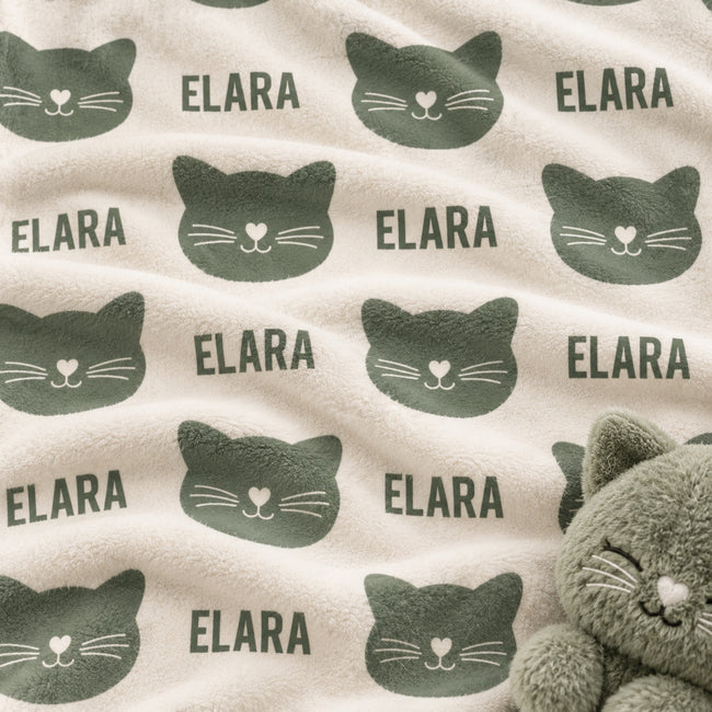 A close-up of a soft fleece blanket with a repeating pattern of dark green cat faces and the word 'ELARA' on a warm cream background. A plush gray cat toy is partially visible in the lower right corner.