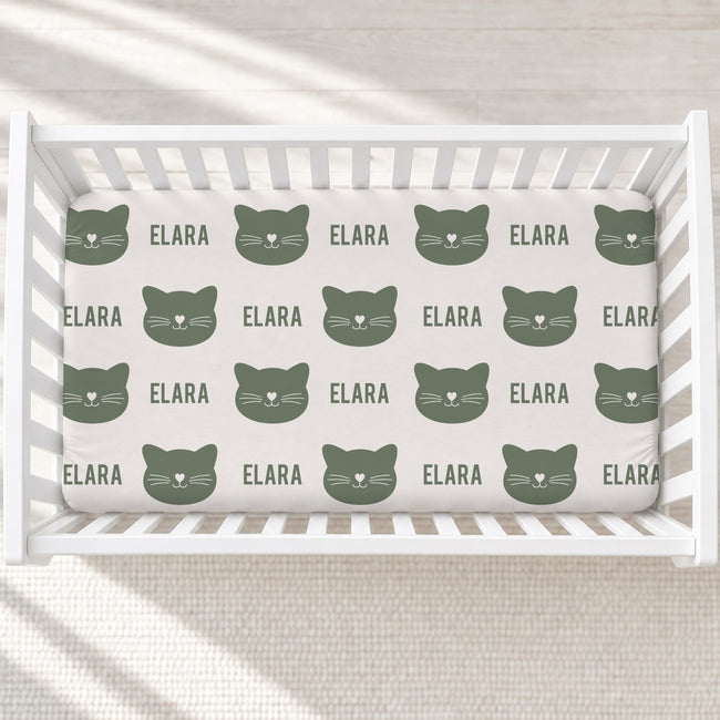 A fitted crib mattress with a repeating pattern of dark green cat faces and the name 'ELARA' in matching green text, shown inside a white wooden crib on a light neutral floor.