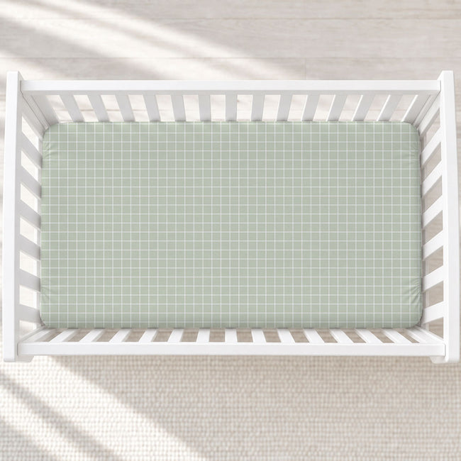 A top-down view of a white wooden baby crib with a sage green and white gingham checkered mattress cover, set against a light beige textured rug with soft diagonal shadows.