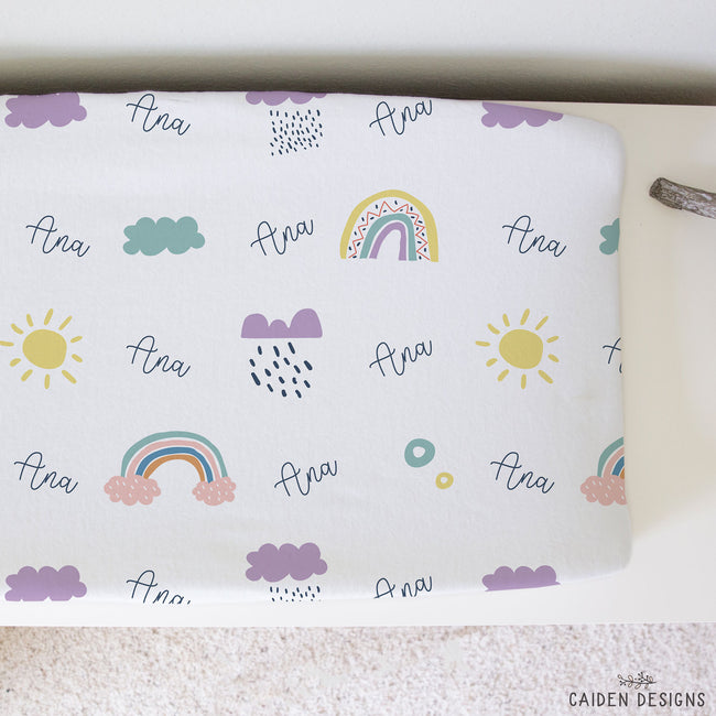 A personalized changing pad cover printed with a whimsical pattern of rainbows, clouds, suns, and the name 'Ana' in script, displayed on a light surface.