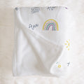A white baby blanket with a colorful rainbow and script text print, laid flat on a white faux fur surface.