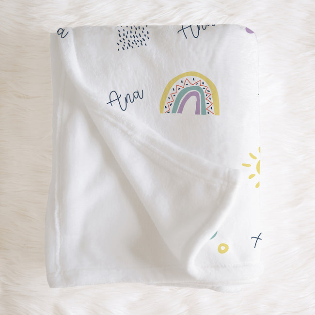 A white baby blanket with a colorful rainbow and script text print, laid flat on a white faux fur surface.