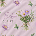 A close-up of a fabric or wallpaper pattern featuring purple and yellow aster flowers, green leaves, and a bouquet tied with a ribbon on a soft pink background. The text 'Evelyn CLAIRE' is printed across the design.
