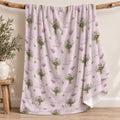 A light pink fleece throw blanket with a repeating pattern of purple aster bouquets and the text 'Emily Claire' is draped over a rustic wooden branch in a styled room setting.