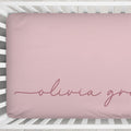 A dusty pink fitted crib sheet with the name 'olivia grace' in cursive script, shown on a mattress inside a white wooden crib.
