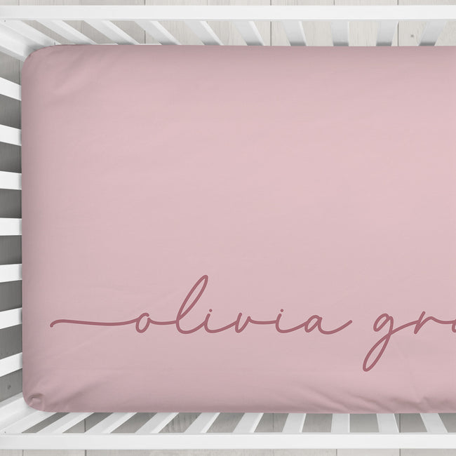 A dusty pink fitted crib sheet with the name 'olivia grace' in cursive script, shown on a mattress inside a white wooden crib.