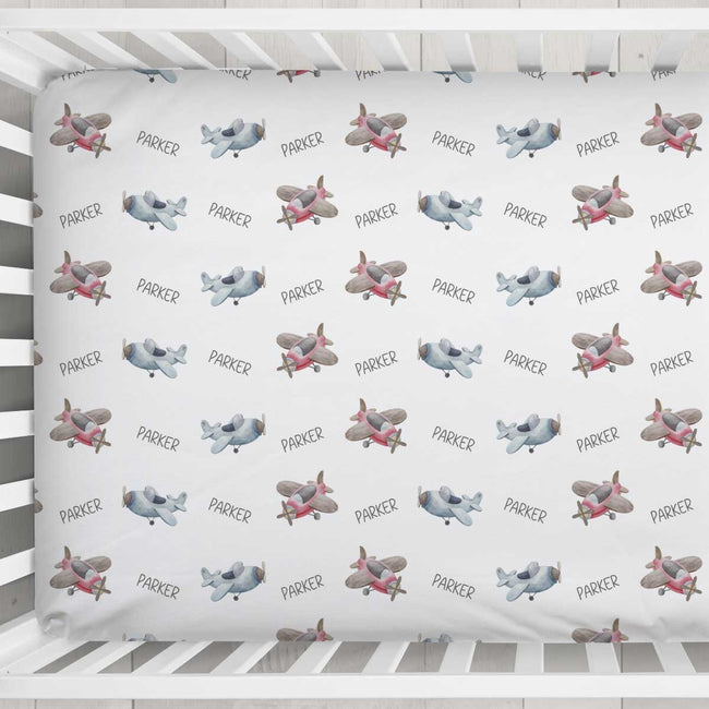 A white crib bedding set featuring a repeating pattern of vintage airplanes in gray, pink, and brown, with the name 'PARKER' printed in black between each aircraft.