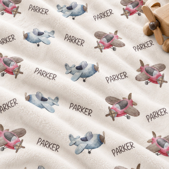 A close-up of a plush cream fabric printed with watercolor-style airplanes in pink, blue, and gray, with the name PARKER repeated in dark gray text. A small wooden toy airplane is partially visible in the top-right corner.