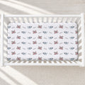 A white crib mattress fitted with a white sheet printed with a repeating pattern of gray airplanes and red rockets, personalized with 'Parker'. The crib is photographed from a top-down perspective on a light beige textured rug with soft natural shadows.
