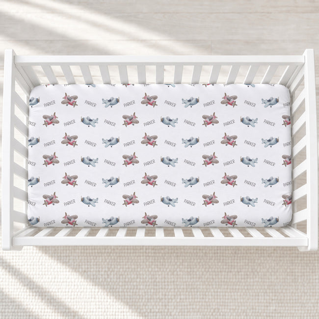 A white crib mattress fitted with a white sheet printed with a repeating pattern of gray airplanes and red rockets, personalized with 'Parker'. The crib is photographed from a top-down perspective on a light beige textured rug with soft natural shadows.