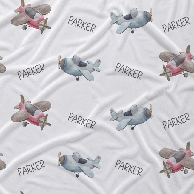 A white fabric blanket or throw featuring a repeating pattern of watercolor-style airplanes and the name 'PARKER' in gray lettering.
