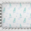 A personalized baby crib mattress cover featuring a repeating pattern of watercolor green dinosaurs, light blue clouds, stars, and hearts, with the name 'Alana' printed in blue script under each motif. The cover is shown fitted inside a white wooden crib.