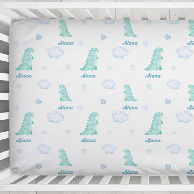 A personalized baby crib mattress cover featuring a repeating pattern of watercolor green dinosaurs, light blue clouds, stars, and hearts, with the name 'Alana' printed in blue script under each motif. The cover is shown fitted inside a white wooden crib.
