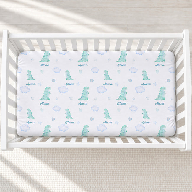 A white crib mattress fitted sheet with a repeating pattern of light blue dinosaurs, hearts, stars, and the name 'Ailana' printed in blue, shown inside a white wooden crib.