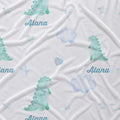 White fabric with a repeating watercolor pattern of light blue clouds, pale blue hearts, and greenish-blue dinosaurs, with the name 'Alana' printed in blue script.