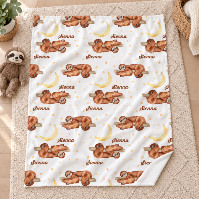 A fleece blanket with a custom print of sleeping brown sloths, yellow crescent moons, and the name 'Sienna' repeated in brown script, laid flat on a beige carpet next to a plush sloth toy.
