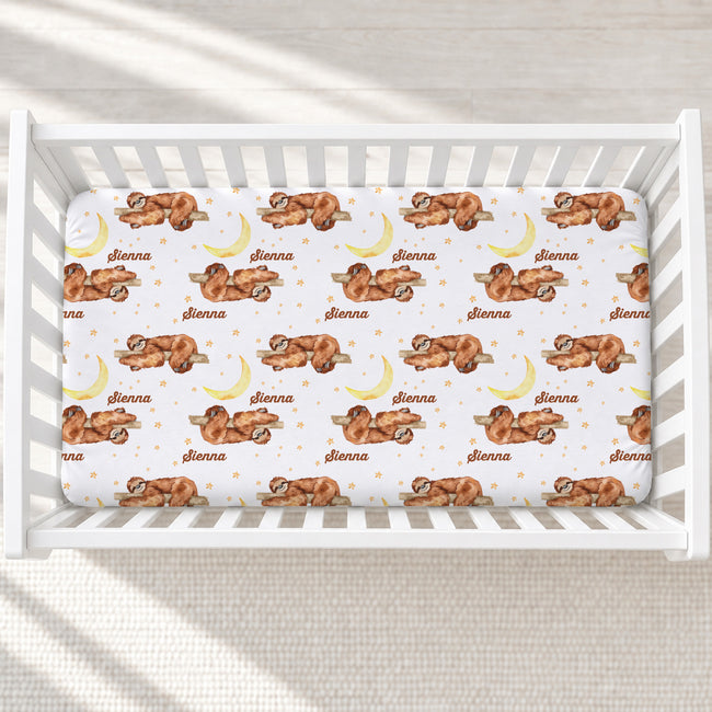 A white crib mattress fitted with a printed sheet featuring a repeating pattern of sleeping sloths, crescent moons, stars, and the name 'shionna' in brown script.