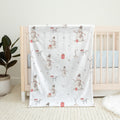 A white baby blanket with a winter snowman pattern draped over a light wood crib in a nursery setting.