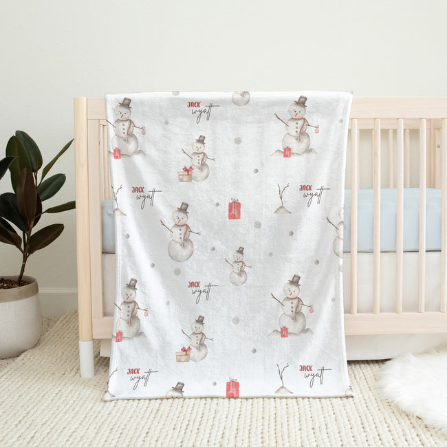 A white baby blanket with a winter snowman pattern draped over a light wood crib in a nursery setting.
