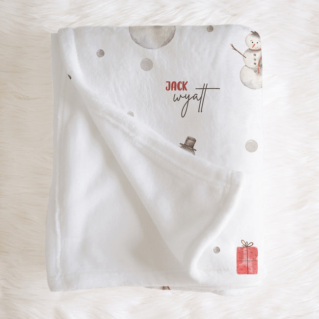 A white personalized baby blanket with a soft fleece reverse side, decorated with a snowman, a small snowman hat, a red gift box, and the custom name 'Jack Wyatt' in red and black script. The blanket is styled flat on a white faux fur background.