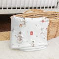 A folded white baby blanket with a winter holiday print of snowmen, gifts, and gray dots, styled on a white faux fur rug next to a woven wicker basket.