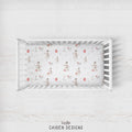 A top-down view of a white wooden crib containing a fitted sheet with a vintage-style snowman print, set against a light whitewashed wood background. The brand name 'CAIDEN DESIGNS' is printed at the bottom.