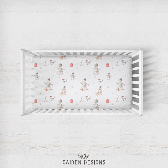 A top-down view of a white wooden crib containing a fitted sheet with a vintage-style snowman print, set against a light whitewashed wood background. The brand name 'CAIDEN DESIGNS' is printed at the bottom.