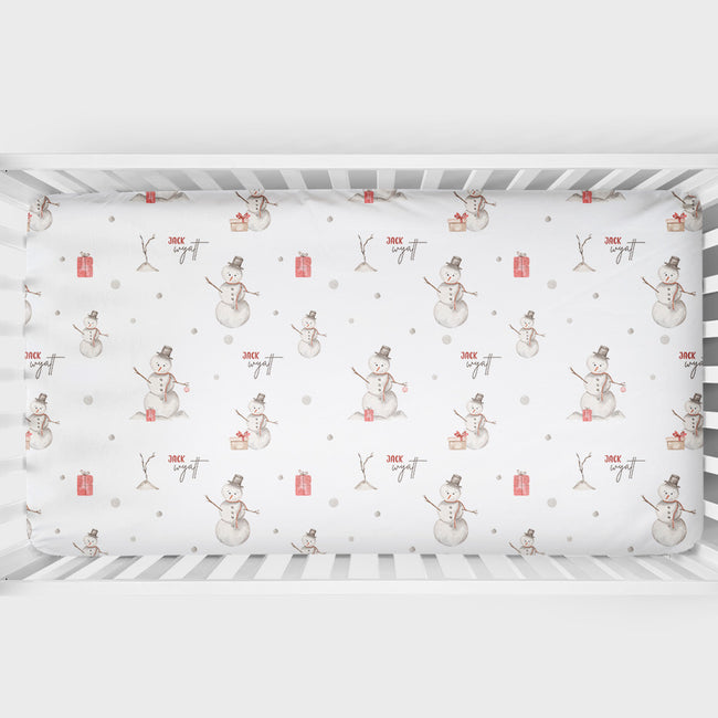 A fitted crib sheet with a winter holiday snowman pattern, shown inside a white wooden baby crib.