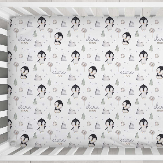 A crib bumper or fitted sheet with a repeating penguin and forest pattern on a white background, shown inside a white wooden crib. The pattern includes penguins, trees, mountains, and the text 'clara mae'.
