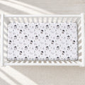 A baby crib mattress fitted with a white sheet featuring a repeating pattern of penguins, trees, and the text 'elara baby'. The crib is shown from a top-down view on a light beige textured floor with soft diagonal shadows.