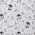 A white fabric blanket with a repeating pattern of cute penguins, pine trees, mountains, and stars, personalized with the name 'clara mae' in gray script.