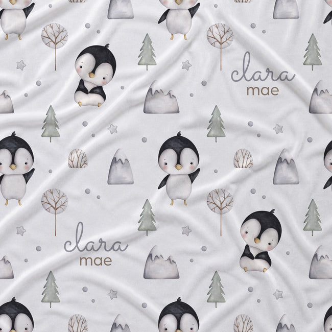 A white fabric blanket with a repeating pattern of cute penguins, pine trees, mountains, and stars, personalized with the name 'clara mae' in gray script.