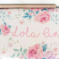 A close-up of a floral-patterned personalized changing pad cover with pink script text reading 'Lola Ann' on a cream background, surrounded by pink roses, blue leaves, and small flowers.
