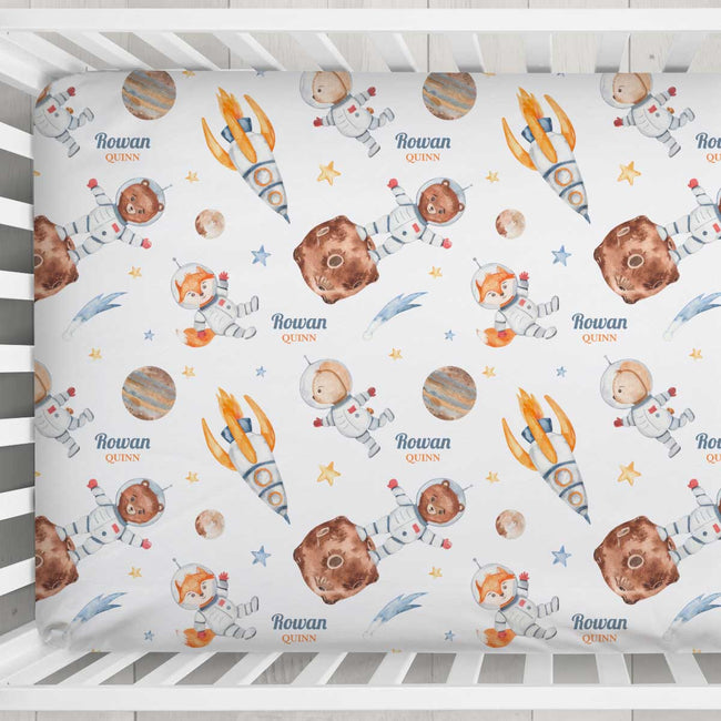 A fitted crib mattress cover with a repeating space-themed print, featuring cartoon astronauts, rockets, planets, stars, and the name 'Rowan QUINN' in blue and orange text, shown inside a white wooden crib.