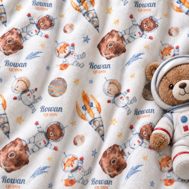 A close-up of a plush blanket or fabric featuring a repeating astronaut teddy bear print with the name 'Rowan Quinn' and small planets/stars. A plush teddy bear wearing a space suit is placed on the right side as a prop.
