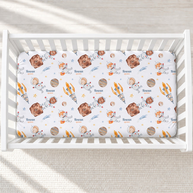 A white crib mattress fitted sheet with a playful space-themed print featuring cartoon astronauts, rockets, planets, and the name 'Rowan' printed repeatedly, shown inside a white wooden crib.