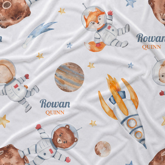 A white fabric blanket or throw printed with a whimsical space-themed pattern featuring watercolor-style astronauts, a fox, a bear, a rocket, planets, stars, and the personalized name 'Rowan Quinn'.