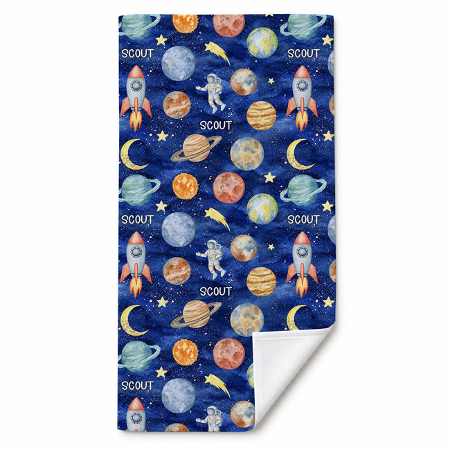 Space Explorers beach towel personalized with Scout, featuring watercolor planets and rockets on navy, with one corner folded to show the white reverse.