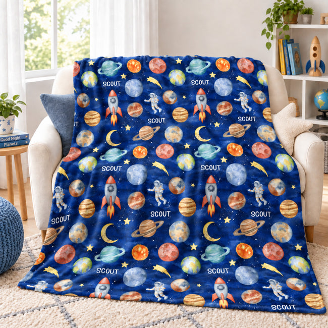 Space Explorers personalized blanket draped over an ivory loveseat, with Scout lettering, watercolor planets, rockets, and astronauts.